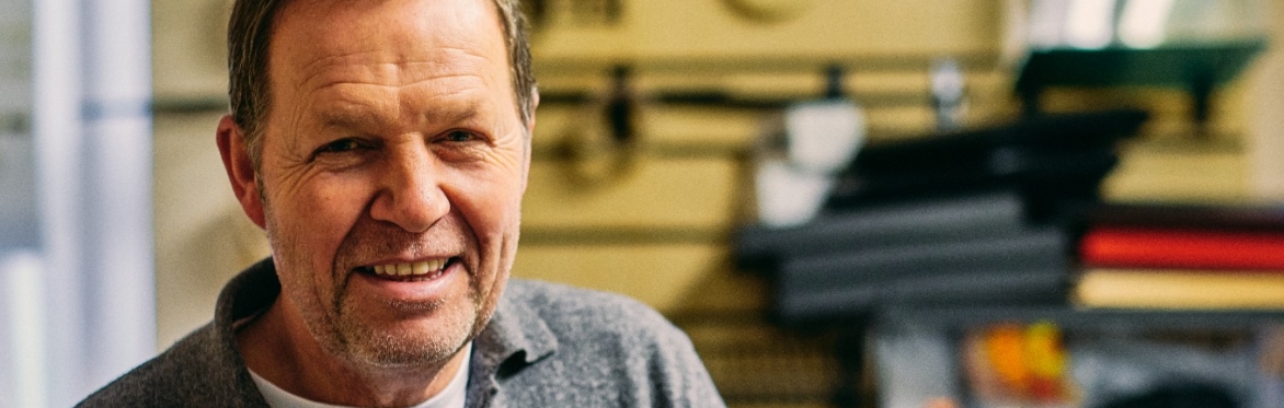 Middle aged male stands smiling to the camera in a workshop. He is wearing a grey jumper.