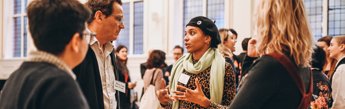 A diverse group of people engage in animated conversation at a networking event. A woman gestures expressively while others listen attentively.