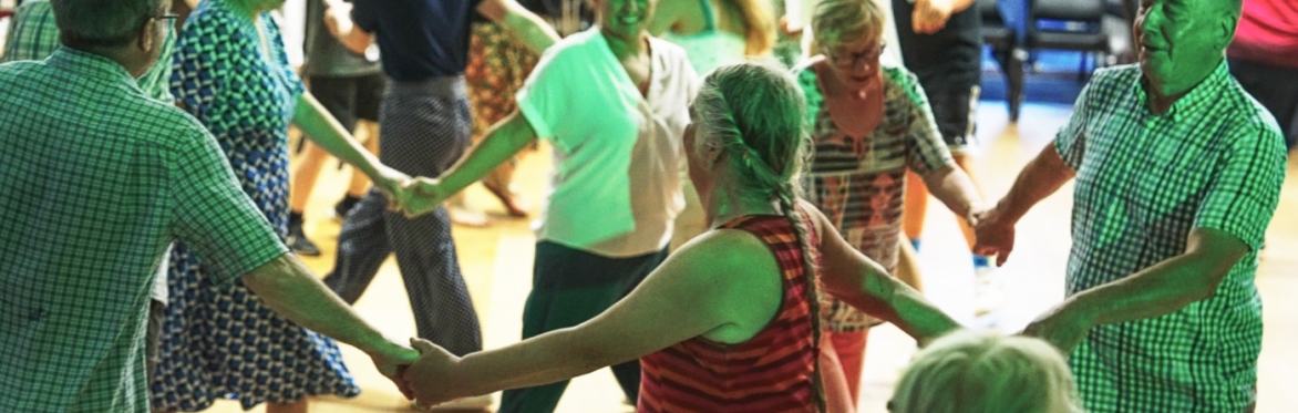 Smiling people of mixed ages and genders dancing together in a circle with their hands joined.
