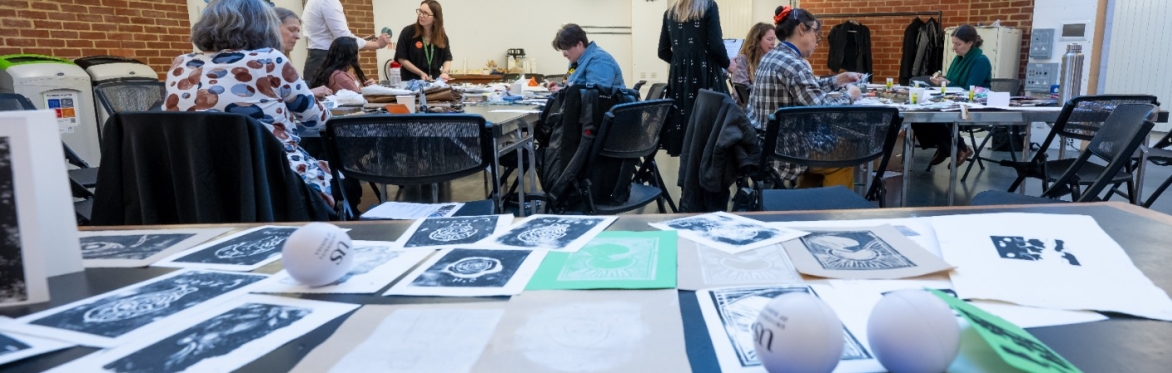 A table of lino prints with people crafting in the background