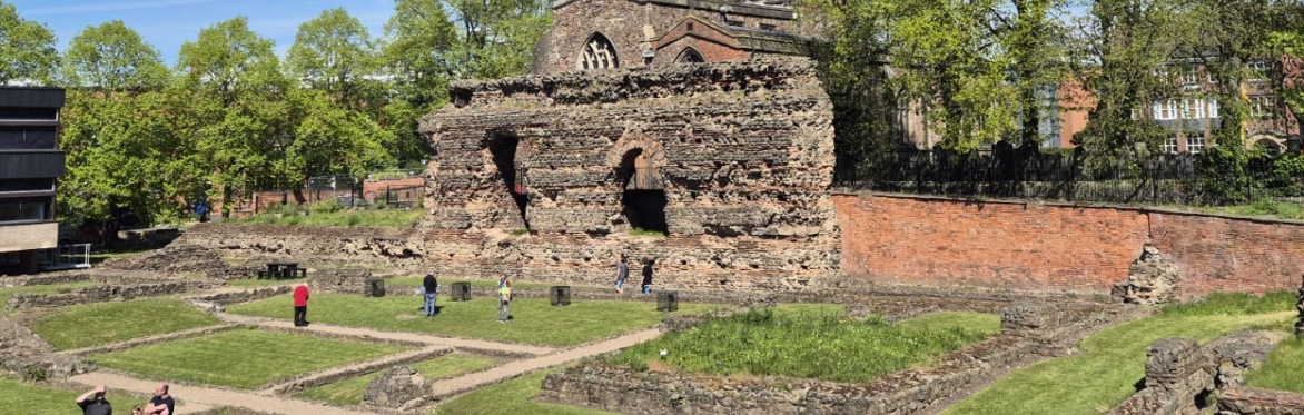 A photo of the Jewry Wall Museum on a sunny day. St Nicholas Church is behind the wall and the rest of the Baths ruins is in front. There are people walking around the ruins.