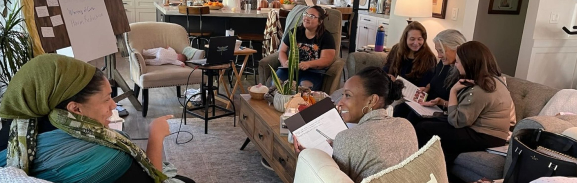 Seven women of different races are sitting in a living room facing an easel. The women are smiling and chatting.
