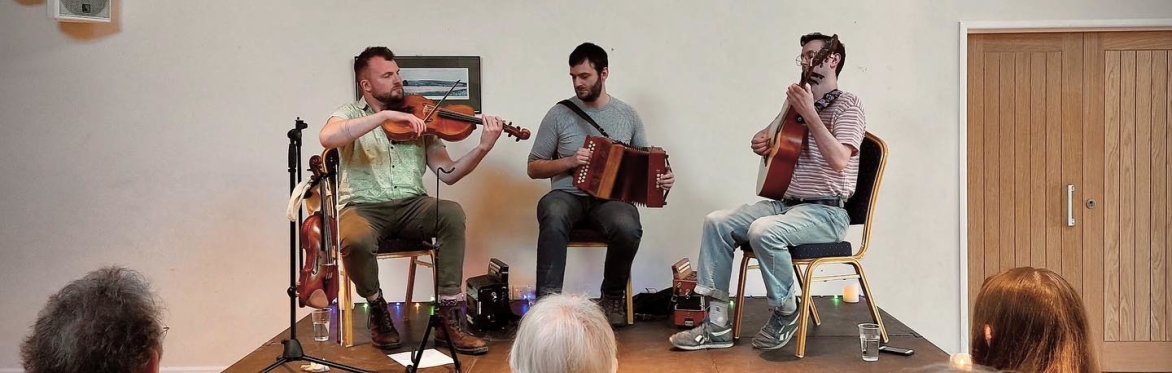 7 members of the band: 5 sitting in a row, 2 standing behind. Some are holding instruments - a saxophone and a diatonic accordian. Behind them are a selection of paintings on the walls.
