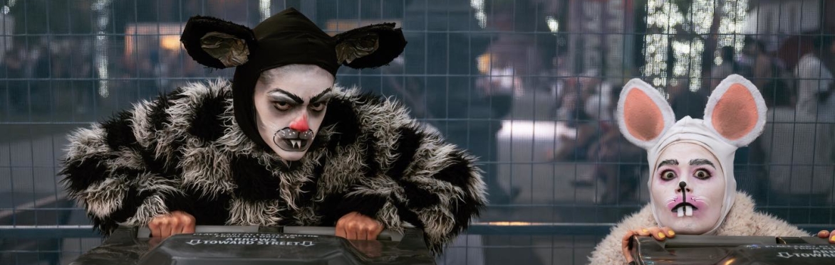 Two people dressed as rats are crouched behind two musical instrument speakers placed on the floor.
