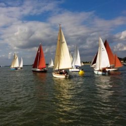 A fleet of traditional sailing boats with white, cream and red sails race across calm estuary waters beneath a dramatic sky filled with large clouds.