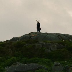 A man with horns and a black coat stands on a rocky outcropping