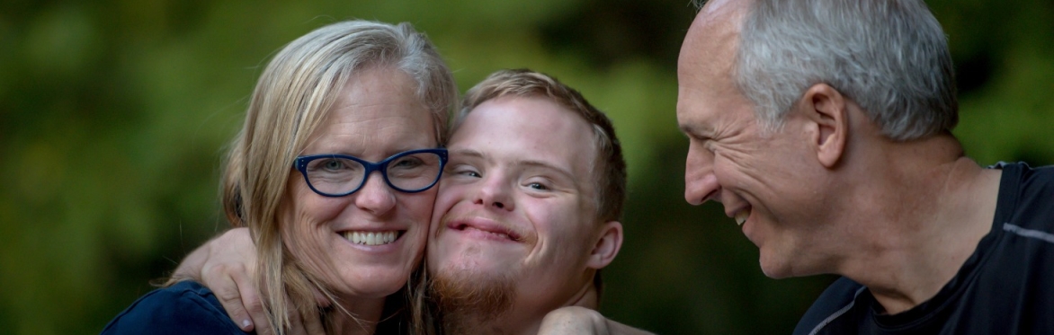 A young man with Downs Syndrome smiling and hugging a woman with blond hair wearing glasses and a white man smiling at them both.