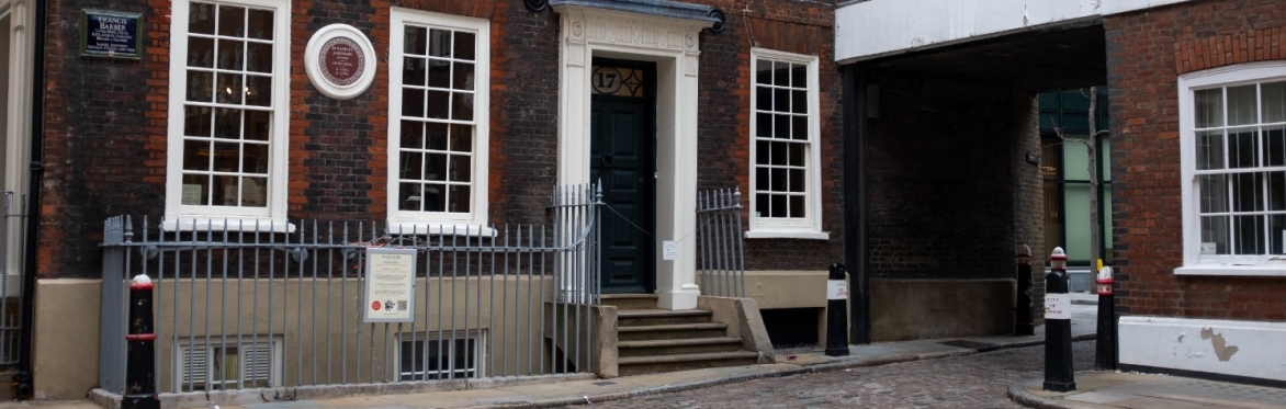 Exterior view of Dr Johnson’s House in Gough Square, London—a Georgian brick townhouse with white-framed sash windows and a dark green front door, accessed by a short flight of steps. A round commemor