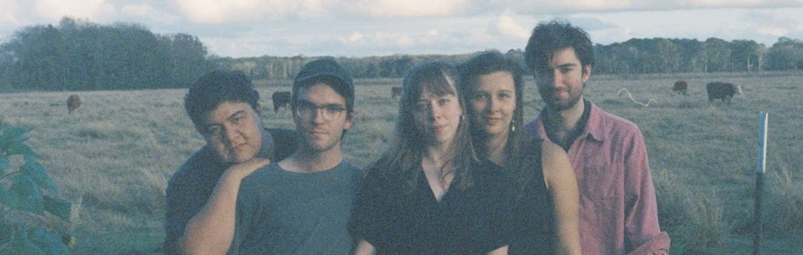 Five members of The Onlies stand closely together in a grassy field at dusk, with cows and a cloudy sky in the background.
