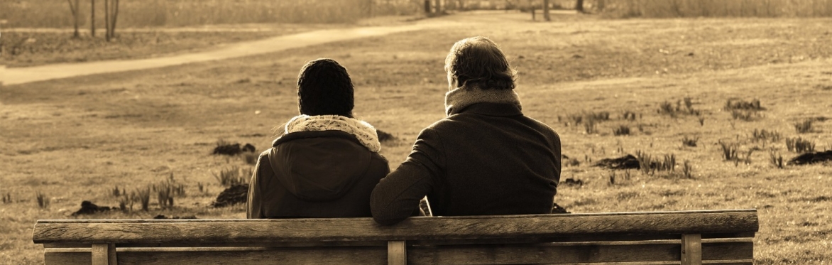 Two people sit next to each other on a park bench, their backs are facing us, they look deep in conversation