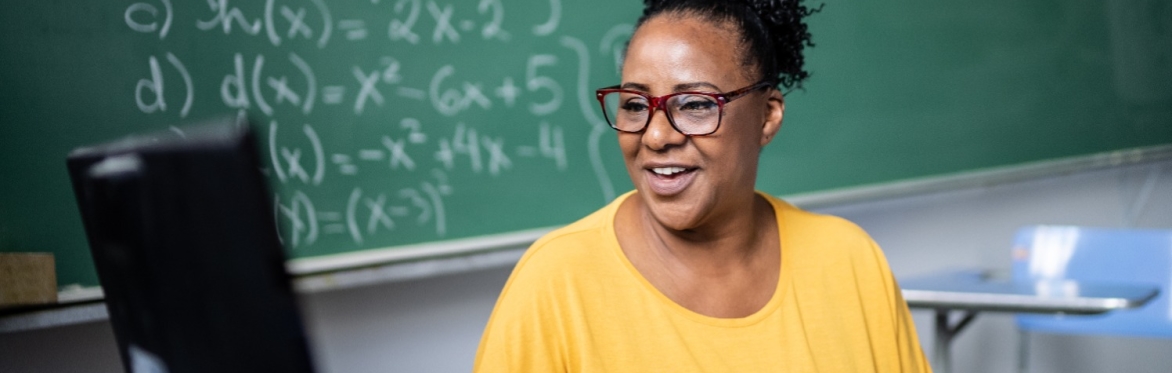 Female teacher sitting at a desk and looking at a computer in front of a blackboard. She has a pencil in her hand and is smiling.