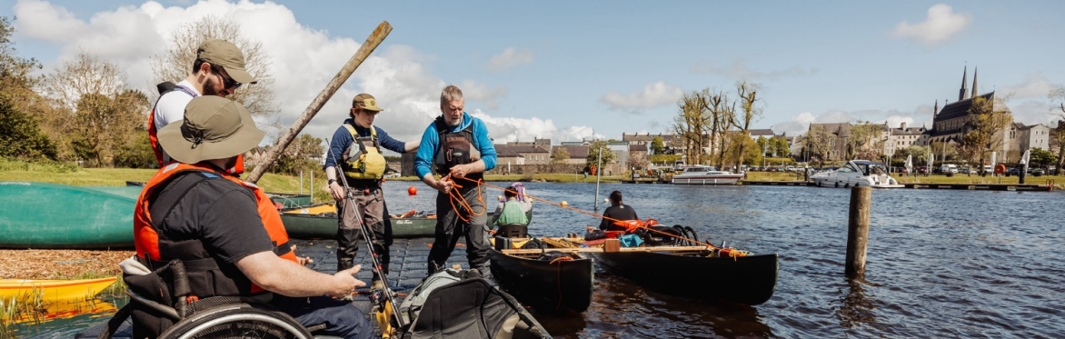 People preparing kayaks and canoes at a riverside dock, including a participant in a wheelchair wearing a life jacket, with others assisting before launching onto the water.