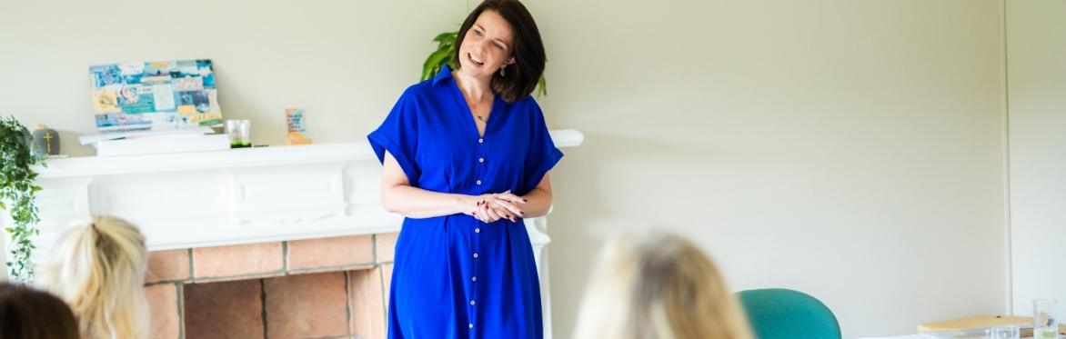 A woman in a blue dress stands in front of a seated group of women working on vision boards