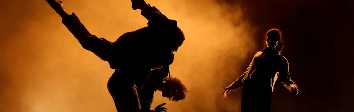 Three contemporary dancers in an improvised moment: one lifting another in motion while a female dancer moves behind them, all set against warm orange light and atmospheric haze