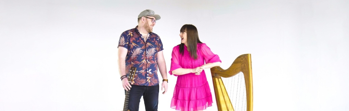 Ron Jappy stands holding guitar with Rachel Hair leaning on her harp