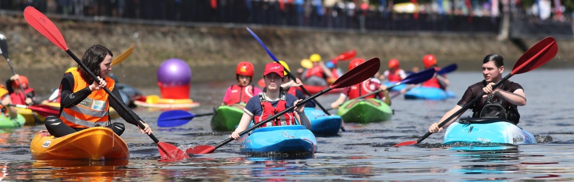 A group of kayakers paddle brightly coloured boats along a calm river, wearing red helmets and life jackets.