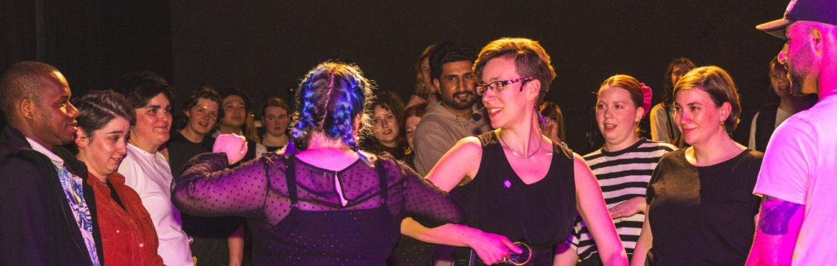 Lisa doing a right arm turn with a person wirh long blue plaited hair and a mid-length rainbow skirt in a dark room with pink lighting