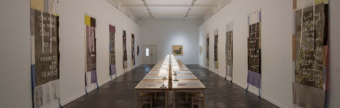 A long table fills a rectangular room. Small white are placed along the length of the table and fabric banners hang on the white walls.