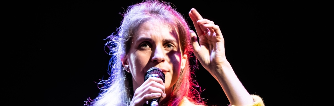 Performer Luisa Muhr stands on stage under dramatic lighting, holding a microphone close to her mouth with one hand and gesturing upward with the other. She wears a textured yellow coat, and her long