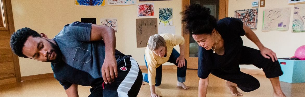 Three dancers of brown, white and tan complexion move low to the ground in matching body shapes, one hand on the floor, knees and one elbow held at right angles, their gazes cast down.