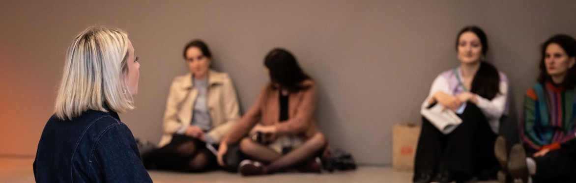 Someone sits on the floor of a gallery, speaking to an audience.