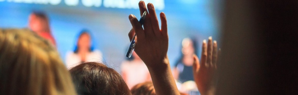 2 people raise their hands at an event. one person has a ben between their fingers. In the blurred background are people a stage