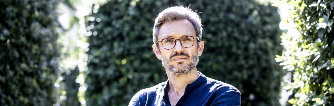 Jason Hickel, a white man with short hair and glasses, stands with his arms crossed. Green hedges and trees can be seen in the background.