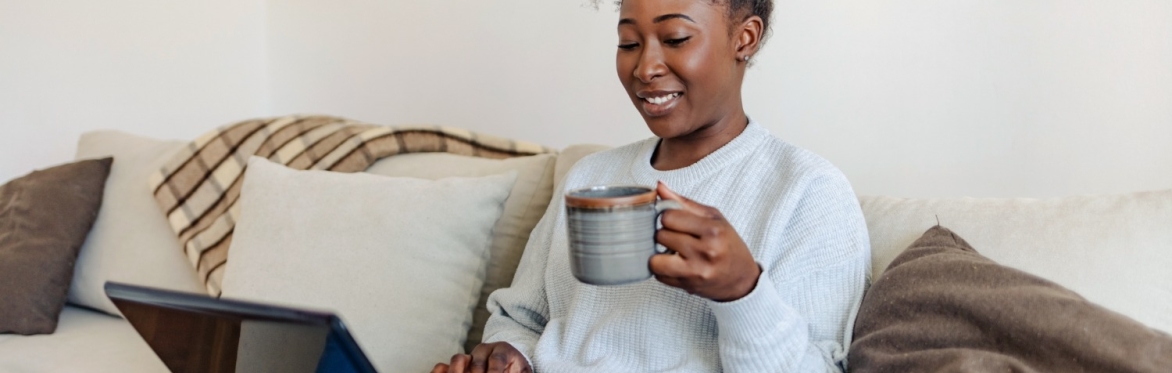 Black woman sat on the sofa using a laptop. She has a cup of tea in her hand.