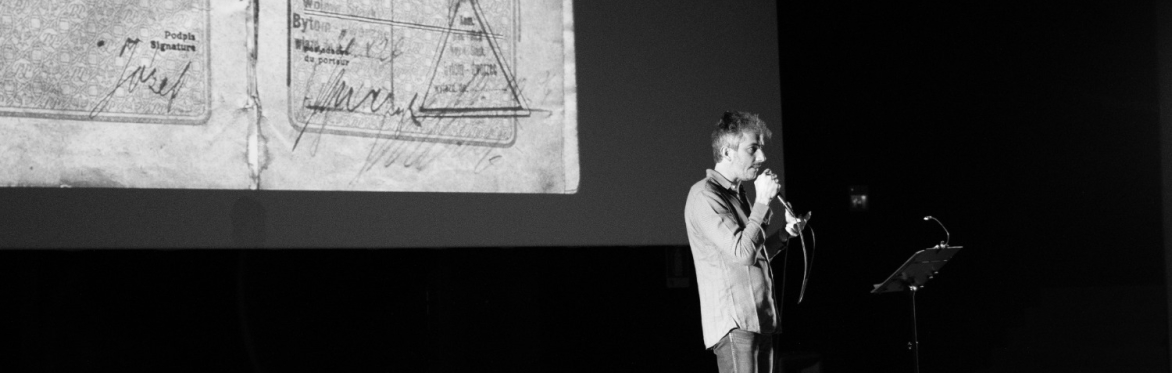 Black and white image of a man speaking into a microphone, stood in front of a screen with a slide of a stamped passport page.