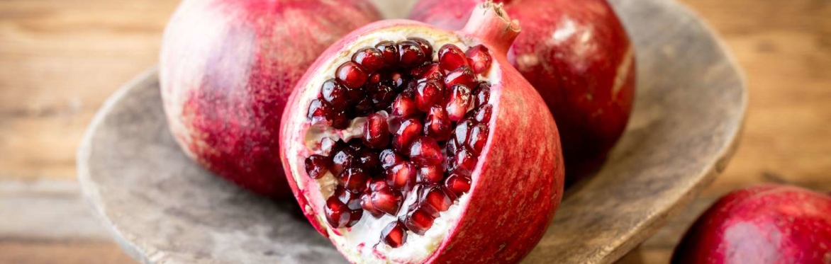 Three pomegranates on a wooden plate. One pomegranate has a wedge removed revealing the seeds inside.