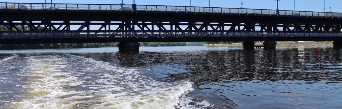 Boat wake on the River Foyle passing beneath the Craigavon Bridge against a clear blue sky.