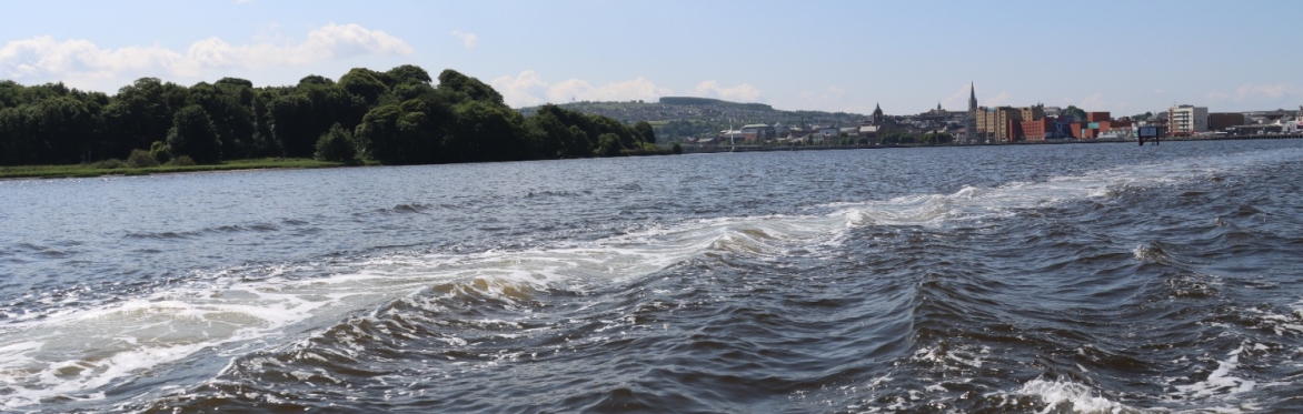 Boat wake rippling across the River Foyle under a clear sky.