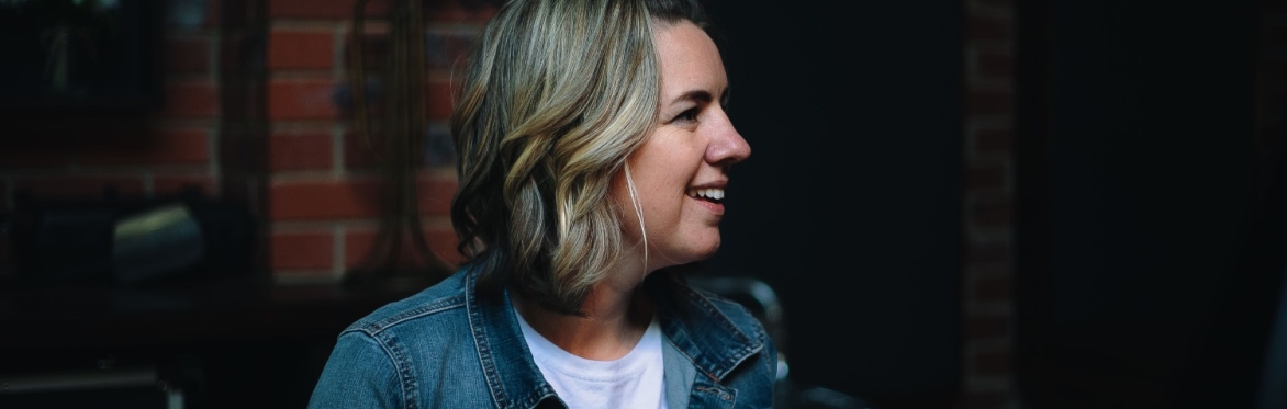 Smiling woman in a denim jacket and white T-shirt sits in profile in a rehearsal space, looking off-camera. Exposed brick walls and music stands create a warm, intimate setting.