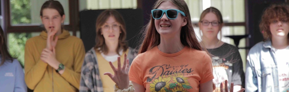 A girl in her late teens stand in front of a group of other young people in a youth theatre workshop. She is wearing sunglasses and waving her arms and  talking