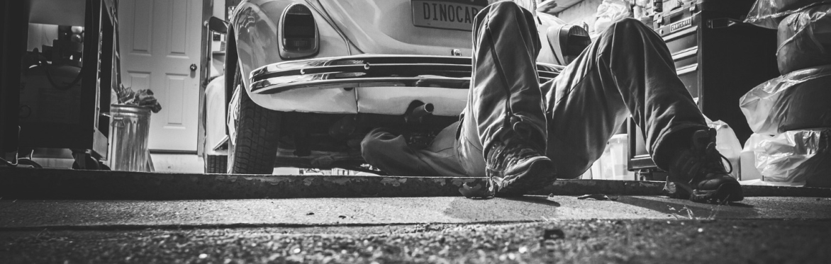 A mechanic lies on their back, looking underneath an older car