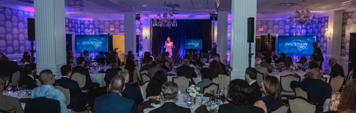 a view from the back of the banquet room of Myra Ruiz welcoming attendees of the 2025 InnoVision awards. Myra is standing on a stage behind a podium. Attendees are seated at round tables.