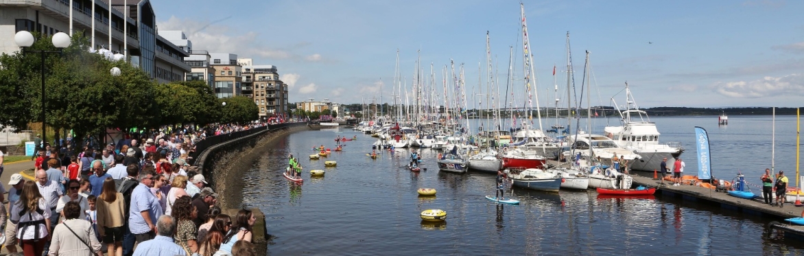 Crowd gathered along a waterfront watching boats and kayakers in a busy marina under a sunny sky.