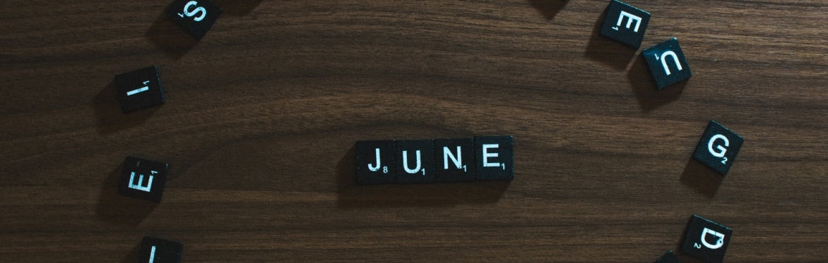 Some dark scrabble tiles in an oval on a wooden table.  4 tiles in the middle of the oval spell the word "June"