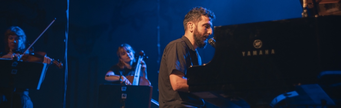 Faraj Suleiman, a Palestinian man, sits at a piano onstage. There is a cello player sitting behind him.