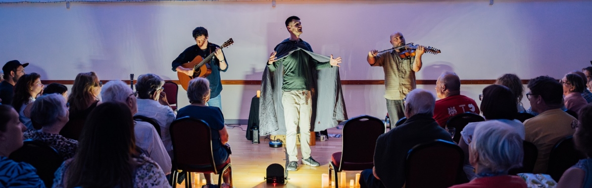 An actor wearing a cloak is speaking in front of a seated audience. The room is lit with atmospheric lights. On either side of him are musicians, one playing a guitar, the other playing a fiddle