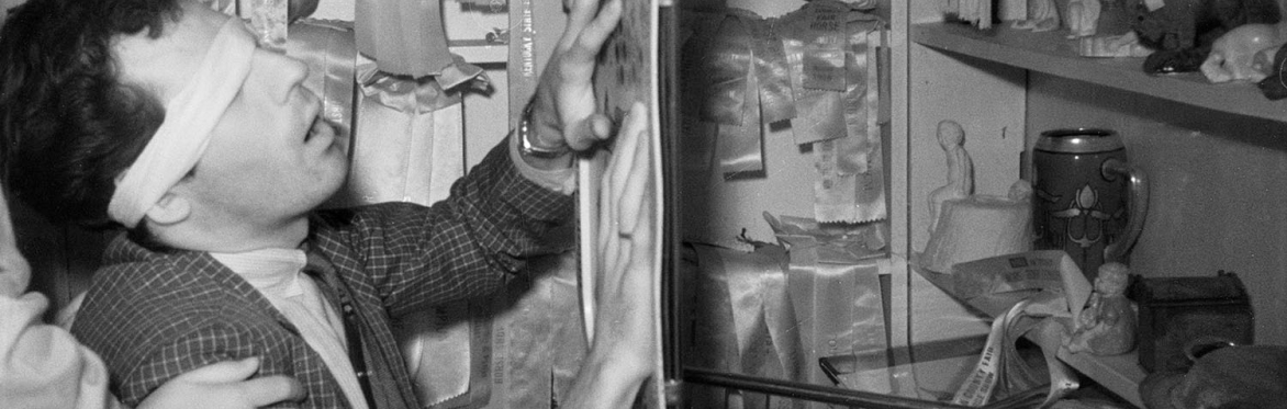 A man wearing a blindfold holds his hands out to touch a small folding table which seems to be suspended in the air