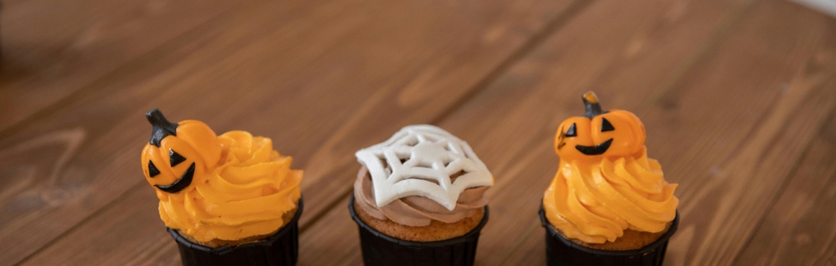 3 Halloween themed cupcakes in a row. The 2 on either end are iced with orange buttercream & topped with a fondant pumpkin. The middle cake has light brown buttercream & a white fondant spiderweb.