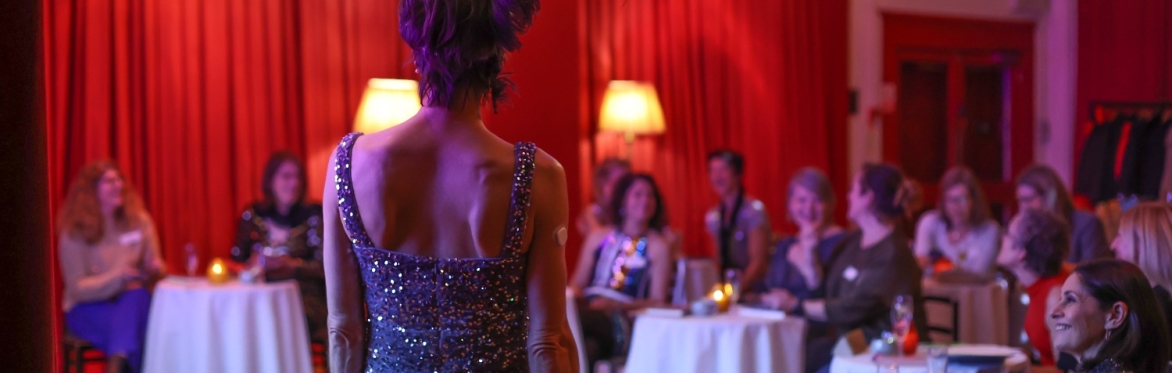 An elegant woman dressed in sequinned dress with a feather in her hair presents investing to a group of women sitting at round tables