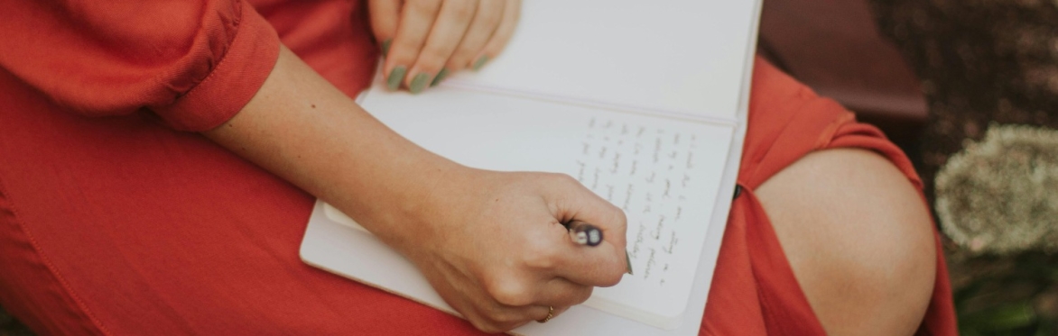 Someone wearing an orange dress, writing in a journal on their lap. Only the person's hands and lap are visible.