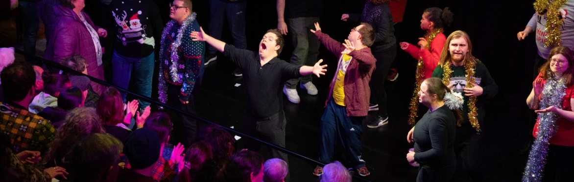A group of performers face a seated audience with arms open wide