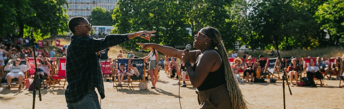 A man and a woman holding microphones sing to each other in front of an audience seated in deckchairs in a park.