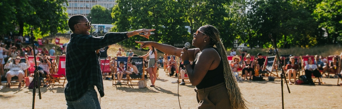 A man and a woman holding microphones sing to each other in front of an audience seated in deckchairs in a park.