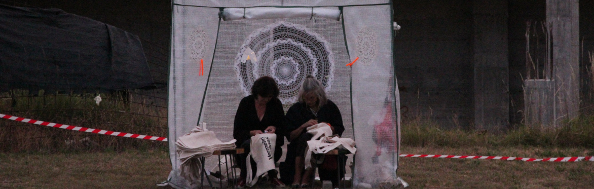 Katrin Woelger und Franziska Helmreich sitzen nebeneinander in einer provisorischen, transparenten Zeltkonstruktion und arbeiten mit Stofftaschen. Hinter ihnen ist ein großes gehäkeltes rundes Textil