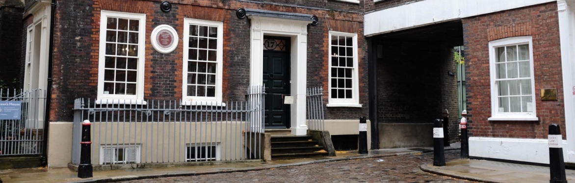 Exterior view of Dr Johnson’s House in Gough Square, London—a Georgian brick townhouse with white-framed sash windows and a dark green front door, accessed by a short flight of steps. A round commemor