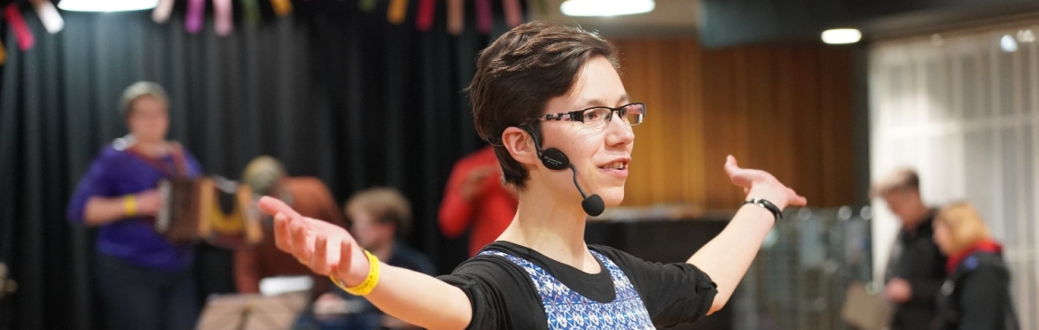 Lisa, a white woman with glasses and short brown hair in her 30s. She is wearing a radio microphone and has her arms open in a welcoming gesture.